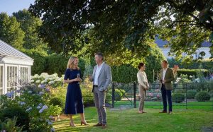 A lush, sun-drenched private garden in late afternoon light, featuring a white-framed conservatory, a black metal estate fence, and vibrant flower beds filled with hydrangeas and roses.